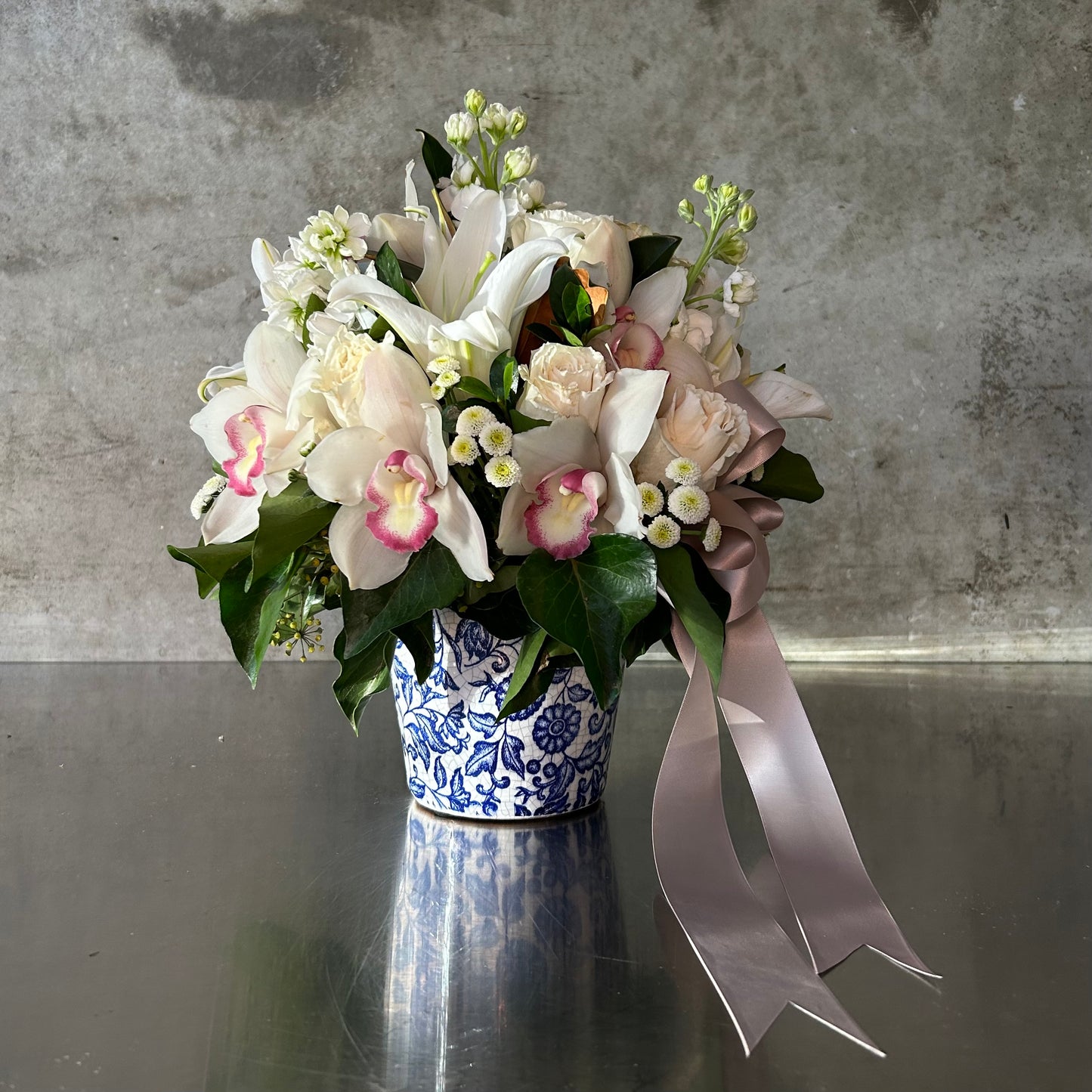Milan soft elegance in porcelain with white lilies, cymbidium orchids, blush roses, petite daisies and trailing greens in a blue floral ceramic pot with taupe ribbon.