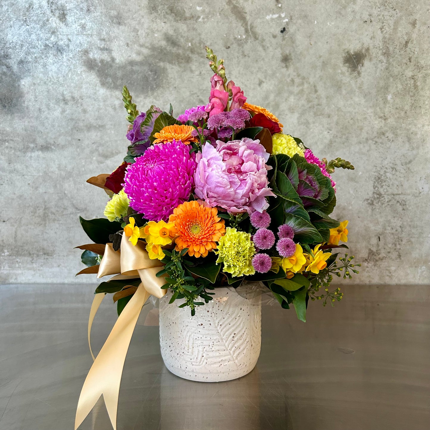 Odesa bold joyful arrangement with vibrant chrysanthemums, peonies, gerberas and seasonal textural blooms in a white ceramic pot with satin ribbon.