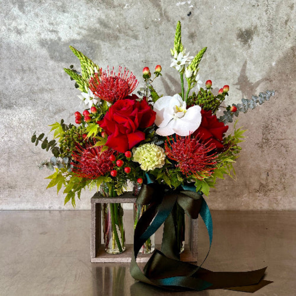 Stockholm modern Christmas arrangement with red roses, hypericum berries, pincushion protea, white orchid blooms, fresh green tones and winter foliage with ribbon finish.