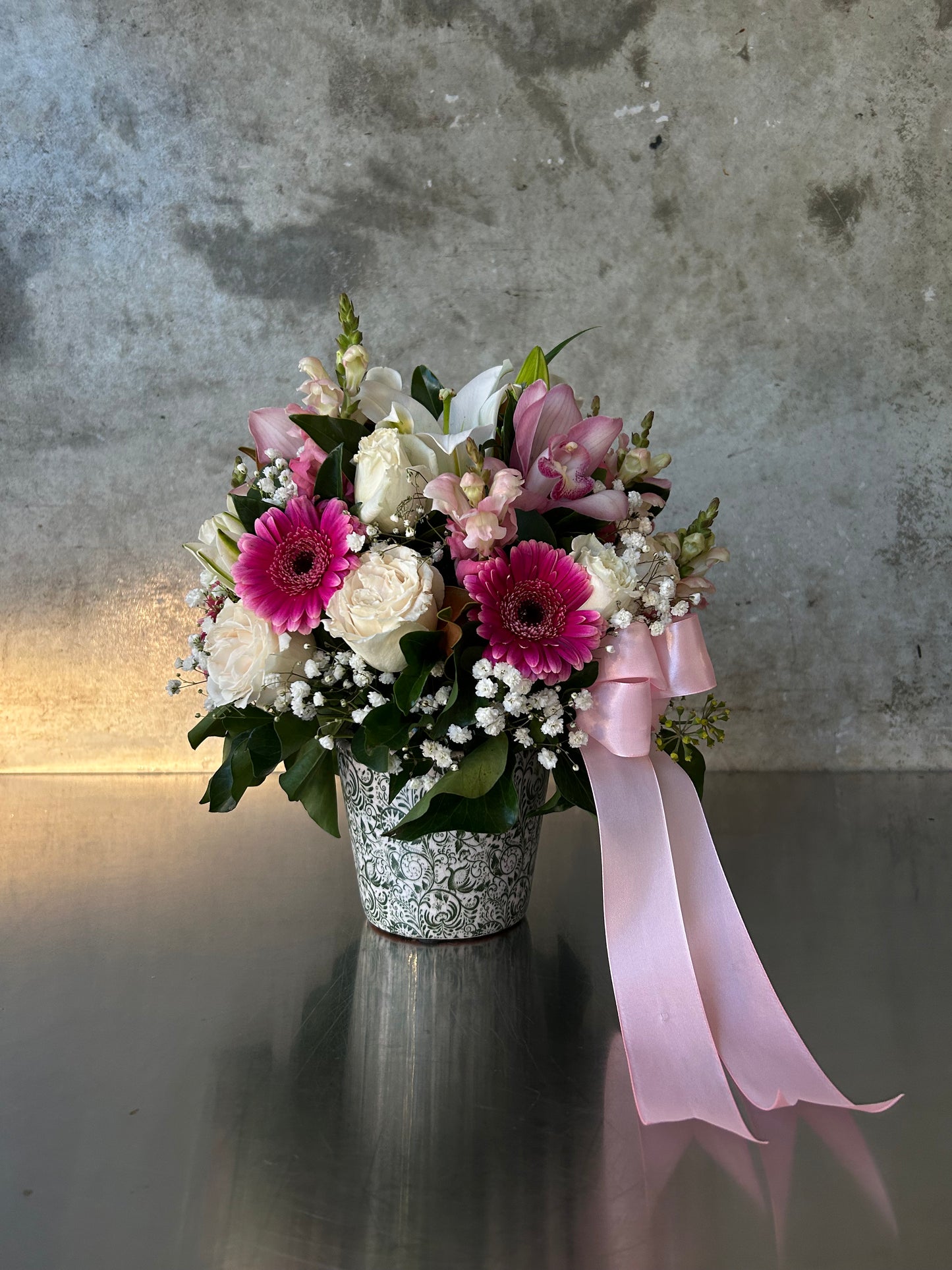 Valencia rose and gerbera garden pot with fuchsia gerberas, creamy roses, snapdragons, soft gypsophila, in a white vessel with pink satin ribbon.